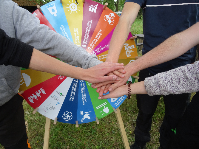 Mehrere Menschen halten ihre Hände zusammen, dahinter sieht man ein Glücksrad mit den 17 Zielen für nachhaltige Entwicklung der Vereinten Nationen